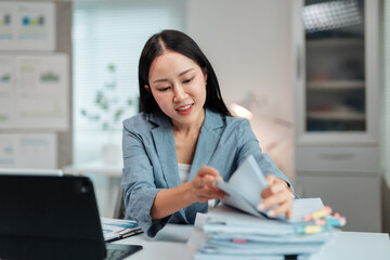 Young Asian businesswoman searching through a mountain of paperwork at her office desk, diligently seeking a specific document among the organized chaos of files and reports
