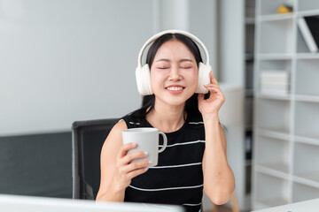 Naklejka premium Young Asian businesswoman is relaxing and enjoying music with headphones while holding a coffee cup during a work break in modern office, representing work life balance and stress relief