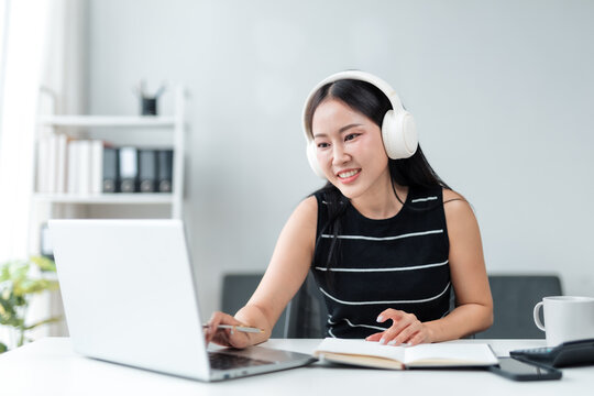 Happy young Asian businesswoman wearing headphones is using laptop computer attending a virtual meeting or online training webinar working remotely from home office - Powered by Adobe