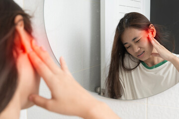 Young woman touching painful ear in mirror with red highlight showing ear infection ache or hearing problem