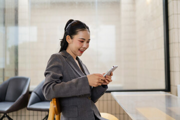 Young Asian businesswoman sitting in a modern office space, smiling and using her mobile phone...