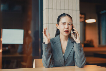 Young Asian manager is having an angry conversation on her smartphone while sitting at her desk in...