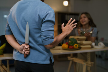 Man hiding a knife behind his back while approaching a woman cooking healthy food in the kitchen,...