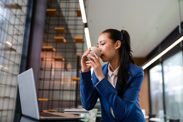 Asian businesswoman in a blue suit sipping coffee or tea while working on a laptop in a modern...