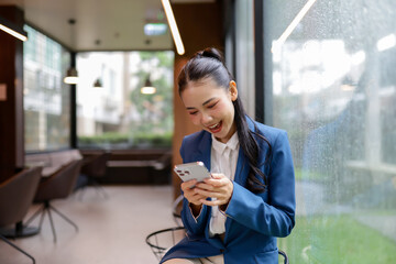 Young Asian businesswoman wearing blue suit using mobile phone and smiling while sitting near window in modern office on a rainy day, enjoying online communication or successful business deal
