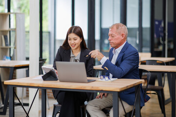 Mature manager mentor talking to Asian female coworker, showing online project results at Business partners in meeting. Two happy diverse professional executives team working on laptop in office 