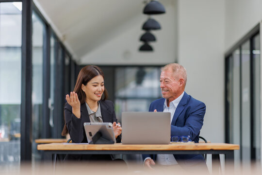 Mature manager mentor talking to Asian female coworker, showing online project results at Business partners in meeting. Two happy diverse professional executives team working on laptop in office 