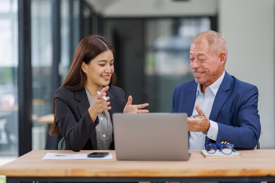 Mature manager mentor talking to Asian female coworker, showing online project results at Business partners in meeting. Two happy diverse professional executives team working on laptop in office - Powered by Adobe