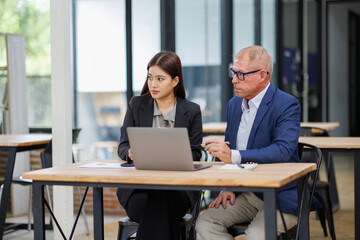 Mature manager mentor talking to Asian female coworker, showing online project results at Business partners in meeting. Two happy diverse professional executives team working on laptop in office 