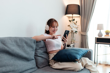 Obraz premium Young Asian woman wearing headphones is using a smartphone and pointing at the screen while sitting on a comfortable sofa in the living room at home