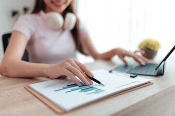 Young professional woman wearing headphones, reviewing marketing data on a clipboard while working on a tablet at her home office desk, focused on analysis and strategy planning