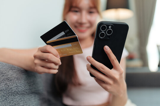 Smiling young woman making an online payment while sitting comfortably on a sofa at home, holding credit cards and using her smartphone for a seamless shopping experience