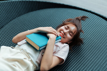 Happy student lying on a round wicker sofa, hugging a book with closed eyes and a joyful smile,...