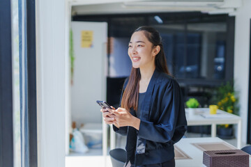 Asian businesswoman is using her smartphone and smiling while looking away in a modern office environment, suggesting a positive and productive work atmosphere