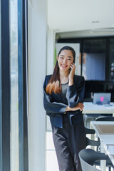 Young Asian businesswoman smiling and chatting on the phone while standing in a modern office near a bright window, embracing her work and enjoying meaningful communication