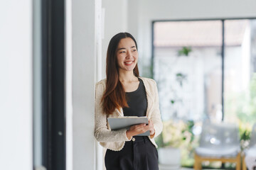 Young Asian businesswoman in smart casual outfit smiling and holding a clipboard while leaning against a wall in the office hallway, looking away