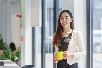 Smiling businesswoman enjoying a coffee break in a modern office, holding a yellow mug and looking towards the camera, embodying the concept of work life balance and relaxation in the workplace