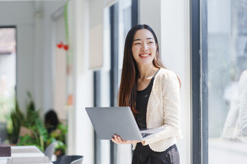 Young Asian businesswoman holding a laptop and smiling while standing by the window in a modern office, enjoying natural light and a positive work environment