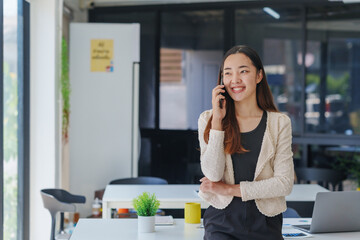 Young Asian businesswoman talking on smartphone and smiling while leaning on desk in modern office, she is wearing a black and white outfit and has long brown hair