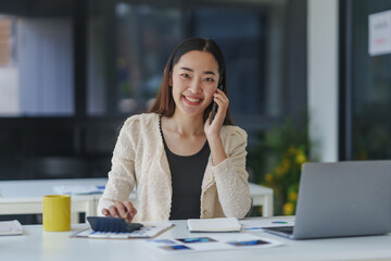 Young professional woman using calculator and talking on the phone while working at her desk in a modern office, managing business finances and multitasking efficiently