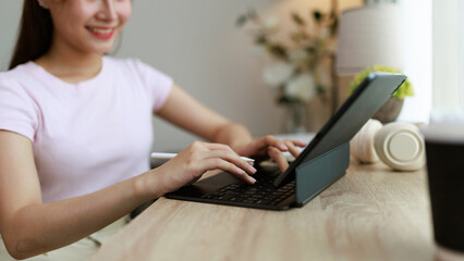 Young woman working from home sitting at a wooden desk typing on a tablet with keyboard and stylus pen, with headphones and a speaker in the background