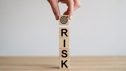 Hand places target block atop stacked wooden blocks spelling RISK, symbolizing business strategy and goal achievement