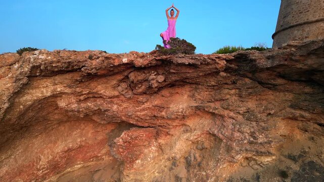 Carefree woman in pink dress Yoga tree pose enjoying freedom and breathtaking view on Ibiza island near Torre des Savinar at sunset. Lovely aerial view flight ascending drone