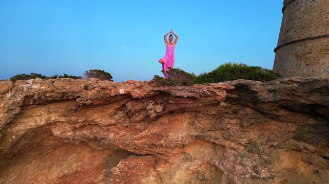 Carefree woman in pink dress with open arms enjoying freedom and breathtaking view on Ibiza island near Torre des Savinar at sunset. ascending drone