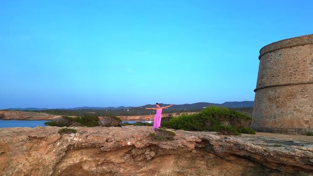 Carefree woman in pink dress with open arms enjoying freedom and breathtaking view on Ibiza island near Torre des Savinar at sunset. Dramatic aerial view flight
