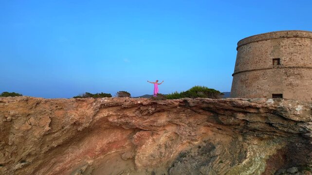 Carefree woman in pink dress with open arms enjoying freedom and breathtaking view on Ibiza island near Torre des Savinar at sunset. Magic aerial view flight descending drone