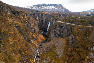 Magnificent Voringsfossen waterfall plunges into deep rugged canyon flanked by rocky cliffs. Winding road and sparse vegetation complete landscape