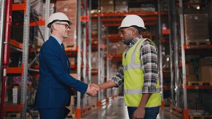 Side view of Caucasian handsome middle-aged male boss wearing hard hat standing in big warehouse shaking hands with young male African American worker, business concept, construction, industrial