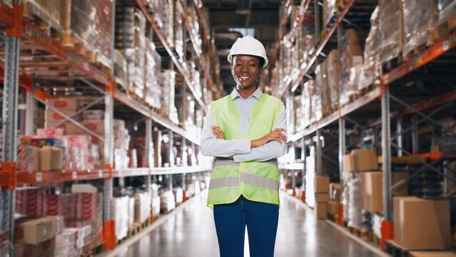 Camera approaching cheerful African American young professional female employee wearing hard hat standing in retail warehouse with many shelves and goods in boxes smiling at camera