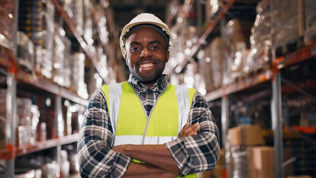 Close up portrait of cheerful African American young professional male employee wearing hard hat standing in retail warehouse with many shelves and goods in boxes smiling at camera, positive mood - Powered by Adobe