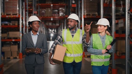 Mixed-race man and women warehouse workers walk through storage racks with merchandise and look up talking in good mood holding tablet and clipboard. Co-workers walking in shelves of logistics factory