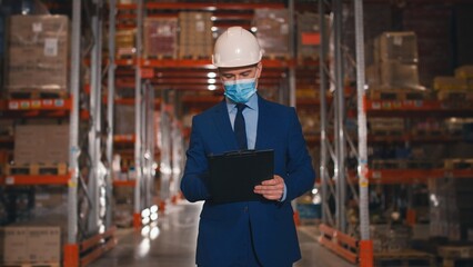 Portrait of Caucasian middle-aged man manager in mask and helmet walk in warehouse with shelves with boxes writing checking inventory. Logistic, distribution center, mixed-race workers on background