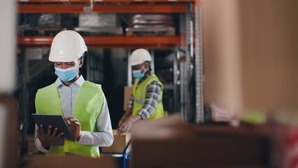African American female worker wearing hard hat and mask on face checks products stock and inventory typing on tablet standing in retail warehouse full of shelves with boxes. Mixed-race people working