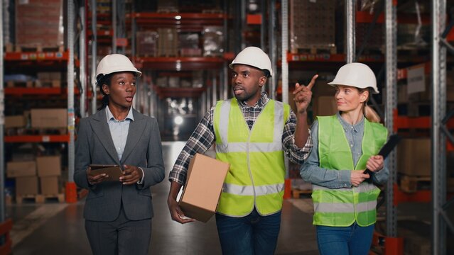 Mixed-race man and women warehouse workers walk through storage racks with merchandise and look up talking in good mood holding tablet and clipboard. Co-workers walking in shelves of logistics factory