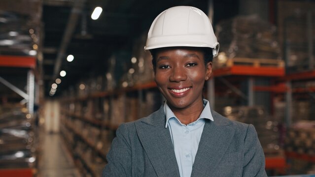 Close up portrait of joyful smiling African American beautiful businesswoman wearing hard hat standing in warehouse and looking at camera. Pretty female worker in helmet with happy face, job concept