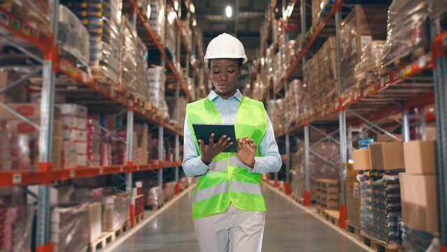 African American young professional woman worker in hard hat uses digital tablet checks inventory walks in retail warehouse full of shelves with goods People working Distribution Center Following shot