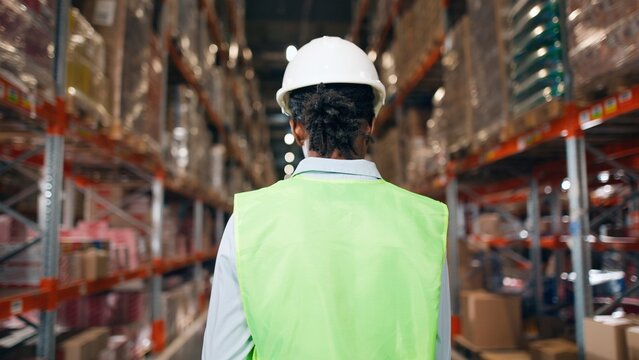 African American young professional woman worker in hard hat uses digital tablet checks inventory walks in retail warehouse full of shelves with goods People working Distribution Center Following shot