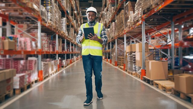 Young handsome African American man using tablet while walking in warehouse of modern factory. Front view of male employee doing work in storehouse holding digital device in hand and looking at screen - Powered by Adobe