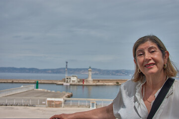 Smiling senior woman poses in front of the iconic port of Marseille under a blue sky with a pier and lighthouse in the background on the coast of France