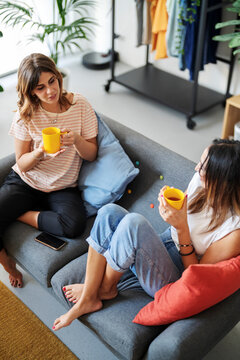 Two women are sitting on a comfortable sofa in a modern living room, holding yellow mugs and engaging in conversation. They are enjoying a relaxed moment together at home.