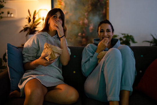Two young women are enjoying a cozy movie night at home. They sit on the couch, eating popcorn from paper bags, and watching something with engaged expressions.