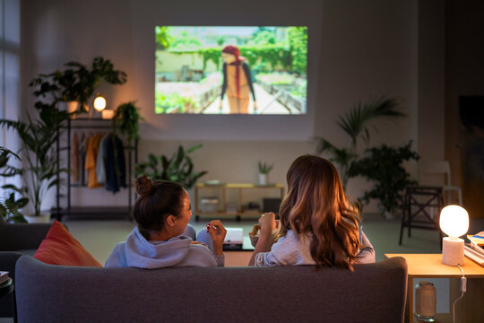 Two women relax on a sofa, watching a movie projected on the wall in a cozy living room. They enjoy snacks during their evening entertainment at home.