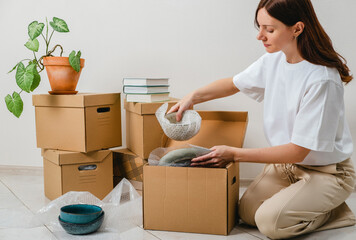 Portrait of woman wearing white t-shirt sitting on the floor surrounded with carton boxes and packing plates and dishes. Relocating, buying new house. Pack personal stuff from carton boxes. 