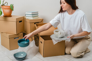 Portrait of woman wearing white t-shirt sitting on the floor surrounded with carton boxes and packing plates and dishes. Relocating, buying new house. Pack personal stuff from carton boxes. 