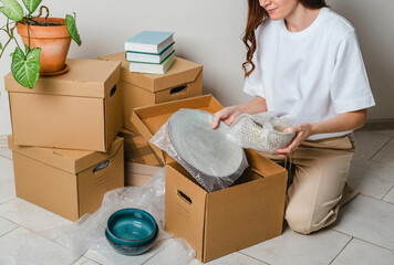 Unrecognized woman wearing white t-shirt sitting on the floor surrounded with carton boxes and packing plates and dishes. Relocating, buying new house. Pack personal stuff from carton boxes. 