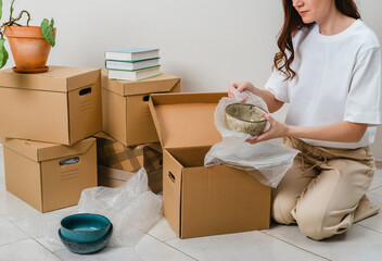 Unrecognized woman wearing white t-shirt sitting on the floor surrounded with carton boxes and packing plates and dishes. Relocating, buying new house. Pack personal stuff from carton boxes. 